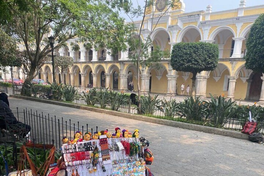 A street with a market stall displaying colorful items and a historic building with arches in the background. 1