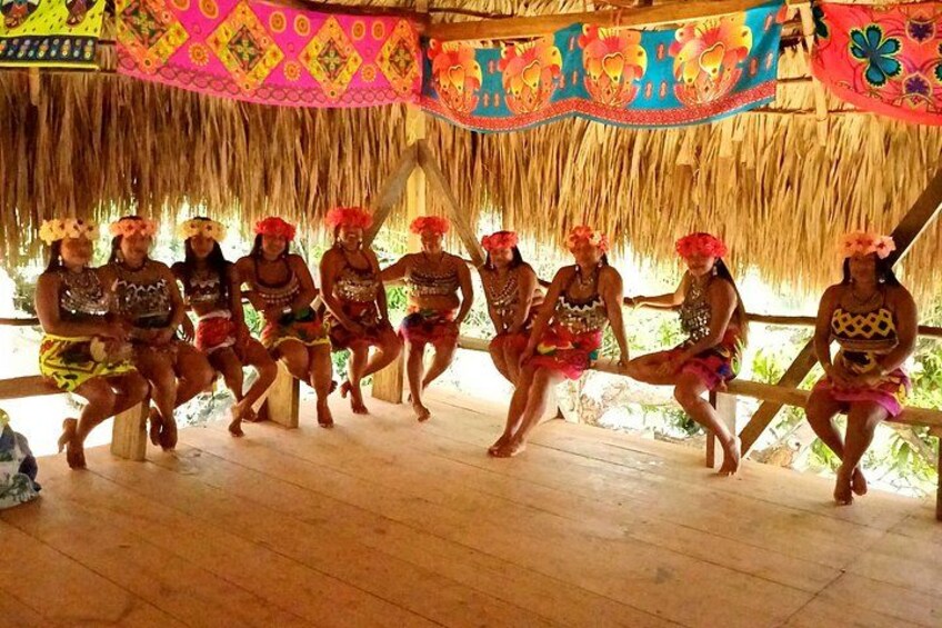 A group of people in traditional attire seated on wooden benches under a thatched roof, with colorful decorations and a tropical setting. 1