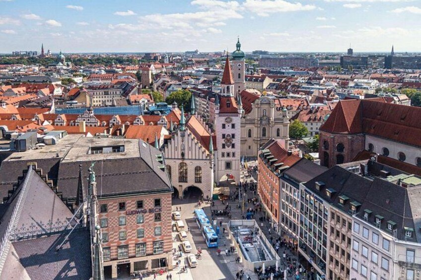 Bybillede med historiske bygninger, en markant kirke med et højt spiretårn og et livligt torv fyldt med fodgængere. 1