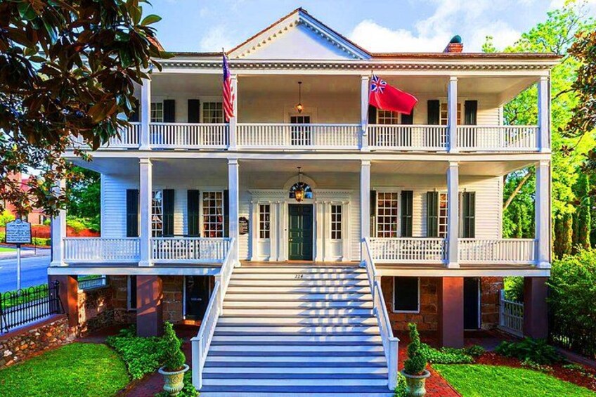 A two-story colonial-style house with a front porch, a flag, and a well-maintained garden. 2