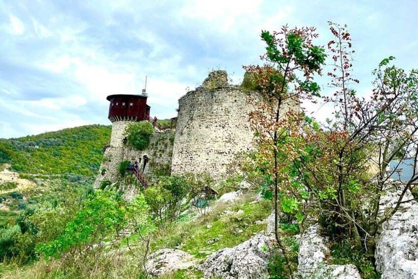 A historic stone fortress with a watchtower, surrounded by greenery and rocky terrain. 1