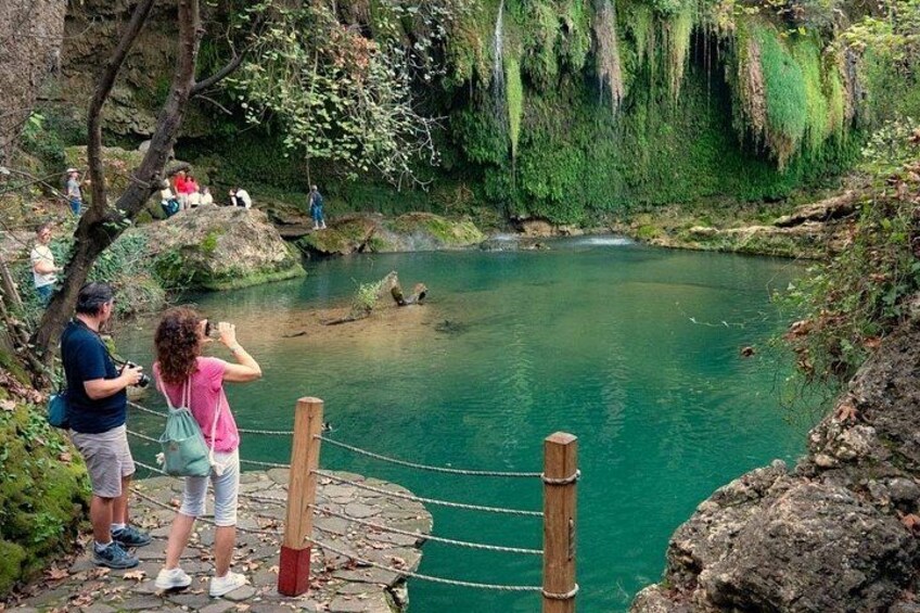 A group of people standing on a wooden platform overlooking a turquoise lake with a rope barrier, surrounded by lush greenery and rocky terrain. 4