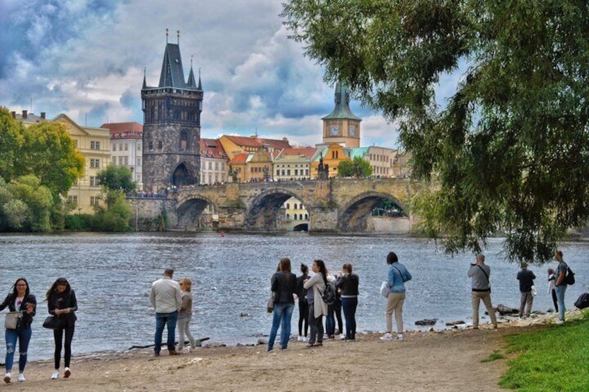 A group of people by a riverside, a bridge, and historic buildings in the background. 2