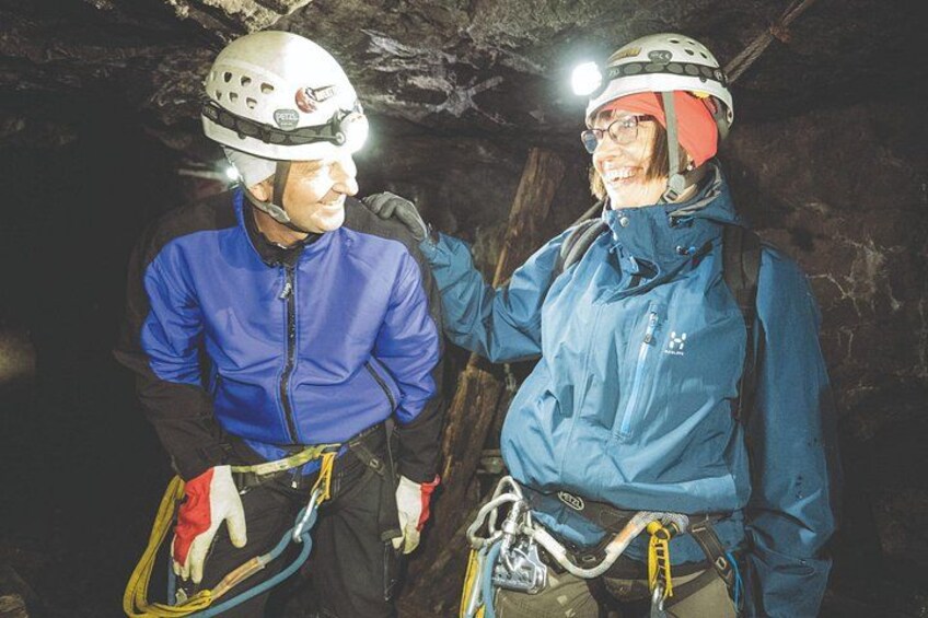 Two people wearing helmets and harnesses, standing in a cave with artificial lighting. 3