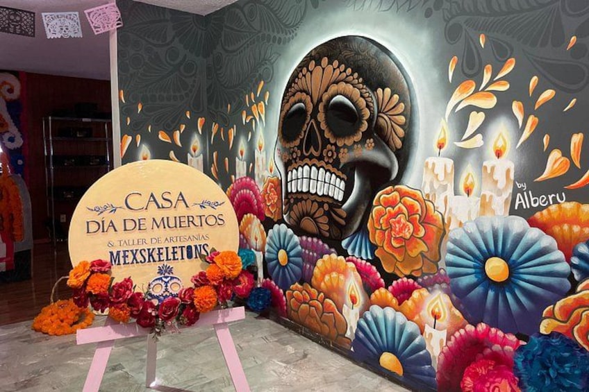 Een ingerichte kamer met een grote muurschildering van een schedel, kleurrijke bloemen en een bordje voor 'Casa de Muertos & Taller de Artesanías Meksikebones'. 5