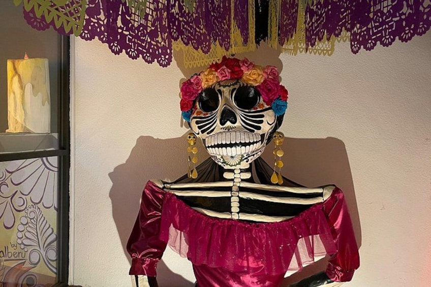Een skeletfiguur met een kleurrijke bloemenkrans en een roze jurk, voor een muur met decoratief papier. 3