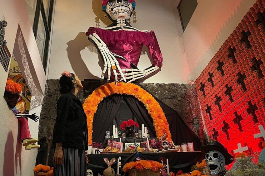 Een skeletfiguur in een paarse jurk, een tafel met aanbiedingen en een rode muur met kruizen. 1