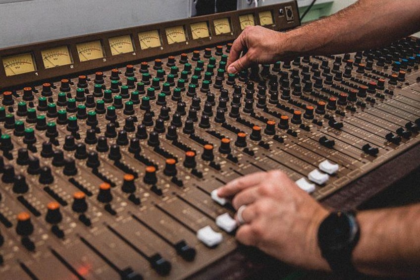 A large mixing console with numerous knobs and sliders, hands adjusting controls, and meters displaying audio levels. 1