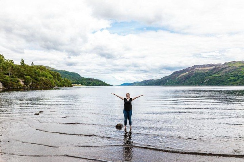 A person standing in shallow water with arms outstretched, surrounded by a calm lake and hills in the background. 2