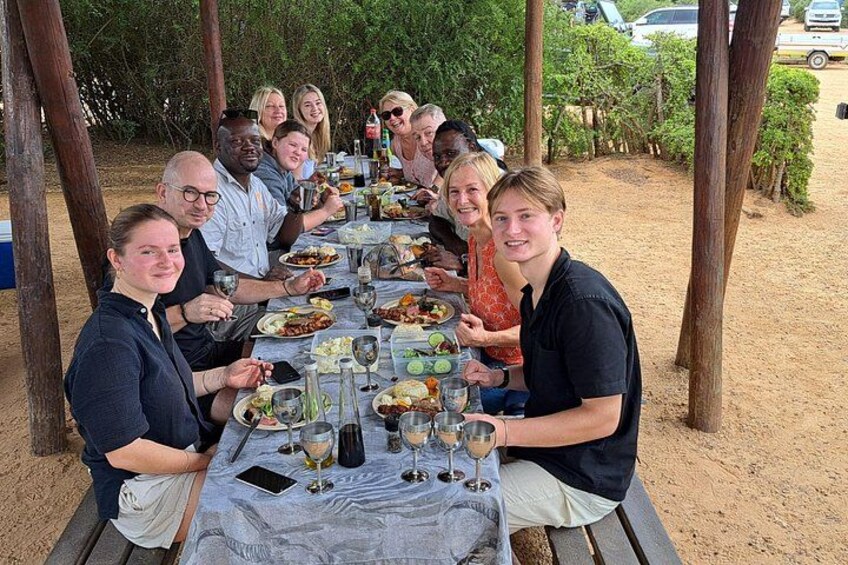 A group of people seated at a long table outdoors, enjoying a meal and drinks. 3
