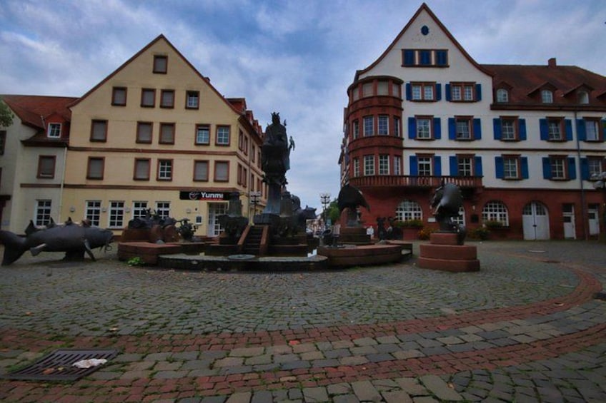 A cobblestone square with buildings, a statue, and a fountain. 1