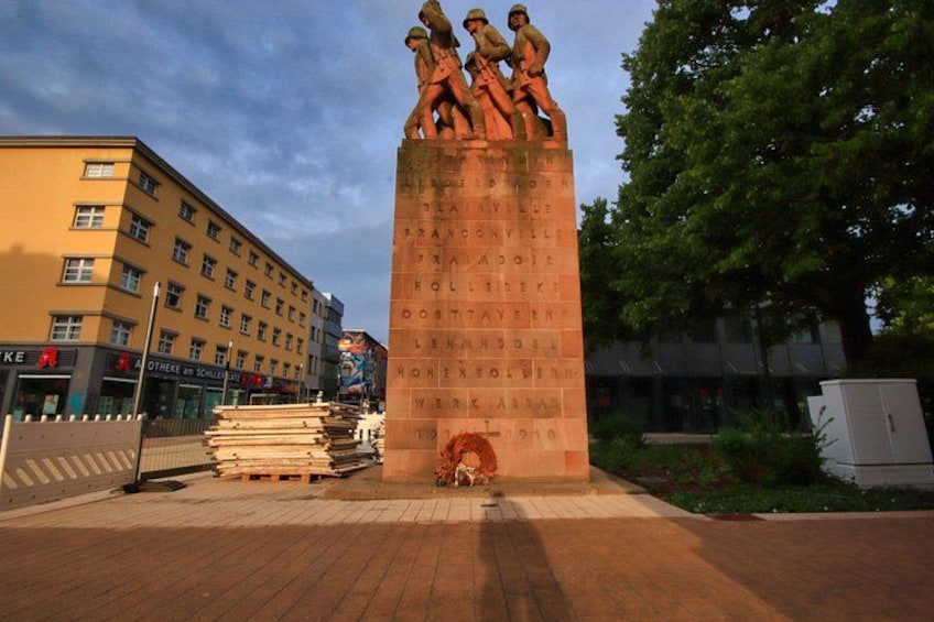 A monument with inscriptions, surrounded by a paved area, with buildings and trees in the background. 5