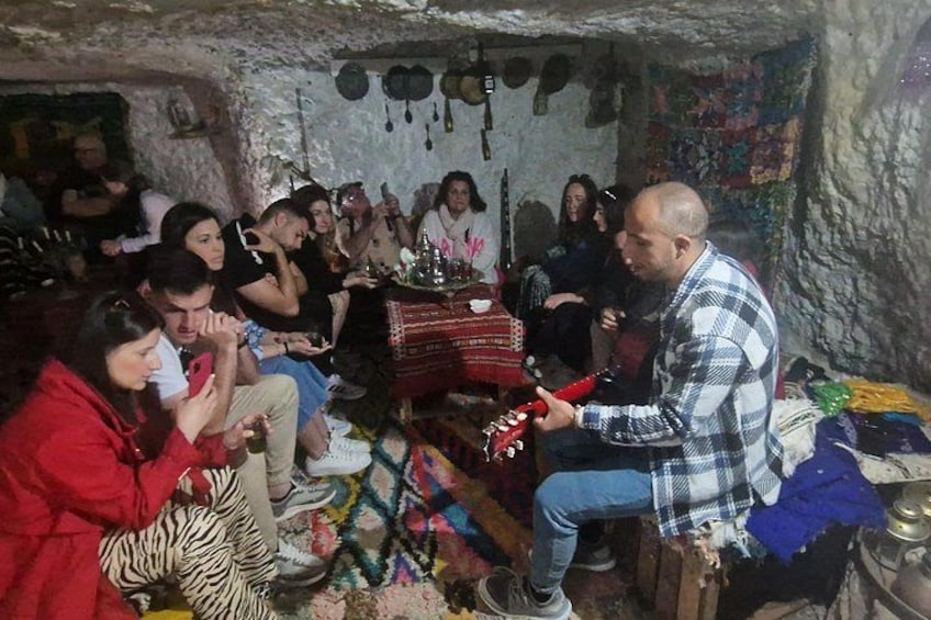 En grupp människor i en grottliknande miljö, några sitter och några står, och en person spelar gitarr. 5