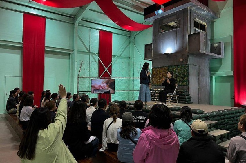 Un grupo de personas sentadas en una sala con cortinas rojas y un escenario donde dos personas hablan. 2