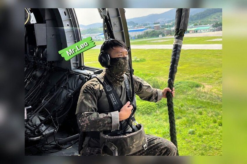 A person in military camouflage attire sitting in a helicopter cockpit with a rope. 5