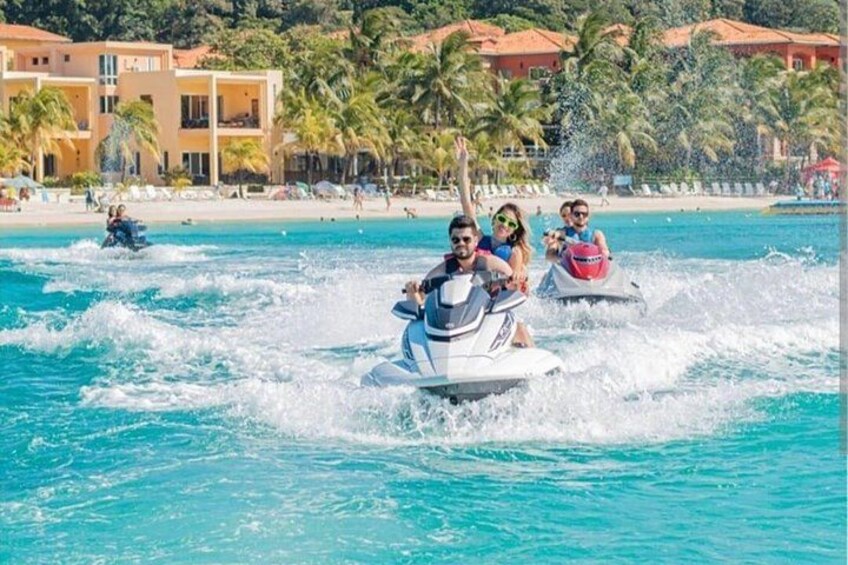 A jet ski with two people and a child on it, water splashing around, palm trees, and a beach with buildings in the background. 4