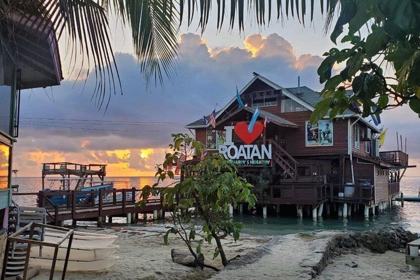A wooden house with a sign reading 'I love Roatan' on a beach with boats and palm trees. 1