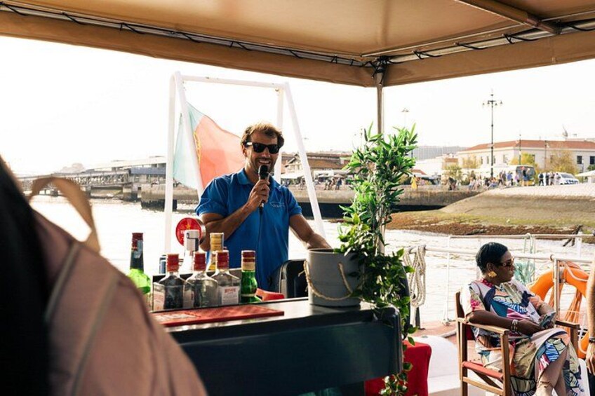 A person standing at a table with bottles and a plant, a woman sitting at a table, and a bridge in the background. 2
