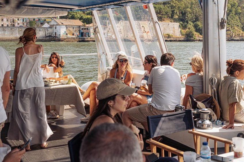 Un groupe de personnes se trouve sur un bateau, autour d’une table dressée pour un repas, avec l’eau tout autour et des bâtiments en arrière-plan. 2