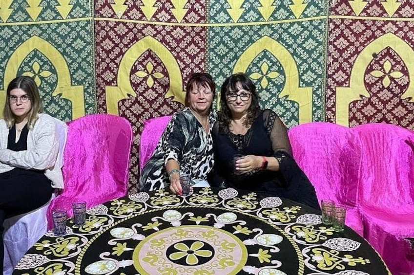 Tre persone sedute a un tavolo con tovaglia fantasia, circondate da sedie rivestite di rosa, con a fare da sfondo un muro decorato con disegni elaborati. 4