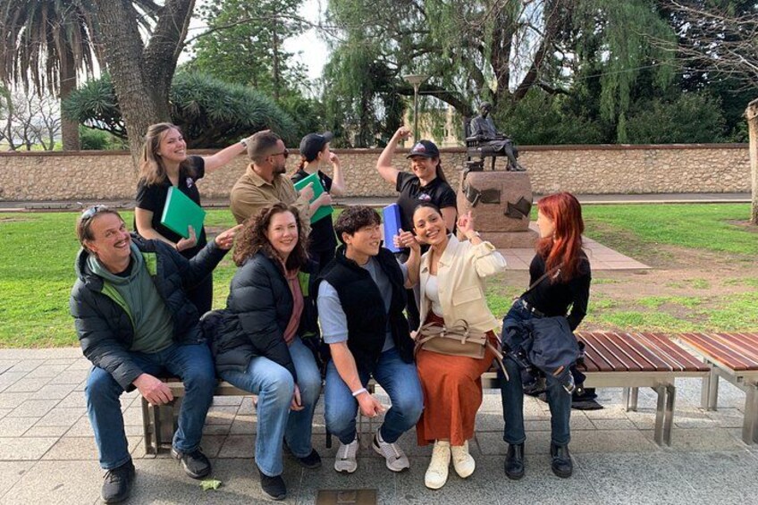A group of people sitting on a bench in a park with trees and a statue in the background. 2
