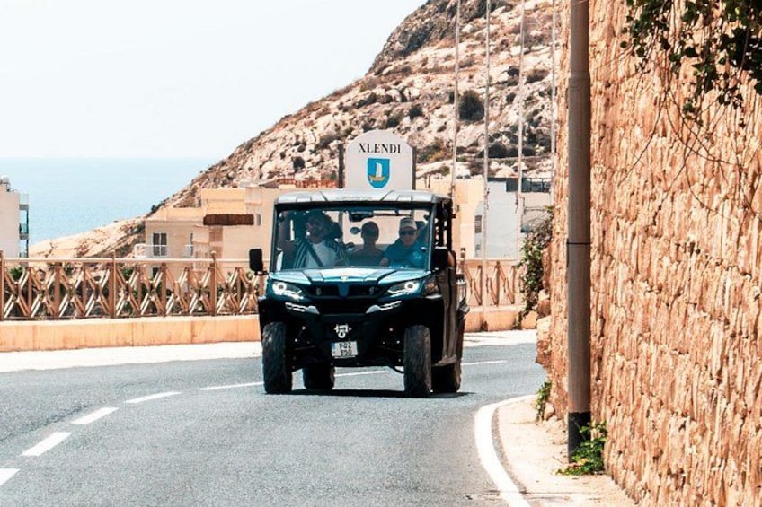 A black off-road vehicle with a sign reading 'XELENDI' on a road with a stone wall and a hilly landscape in the background. 2