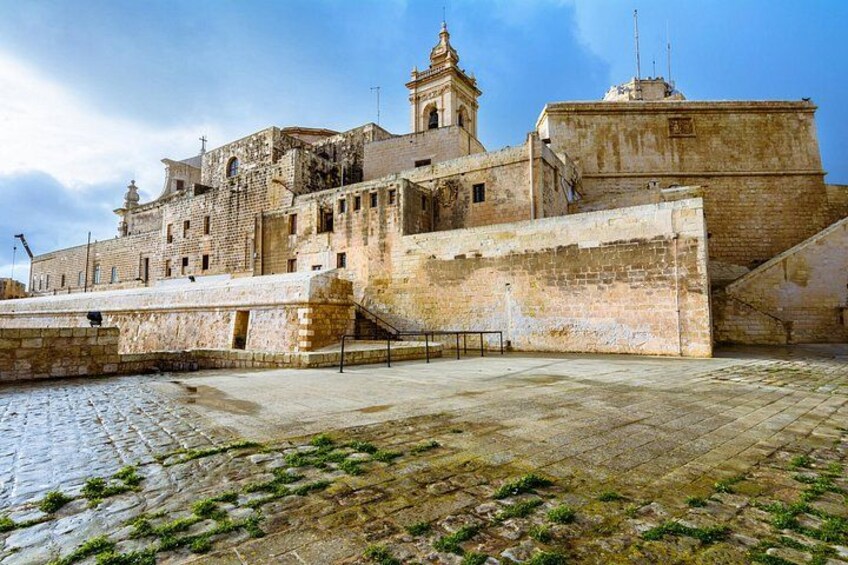 A historic stone fortress with a prominent bell tower, a large open courtyard, and a fortified wall. 3