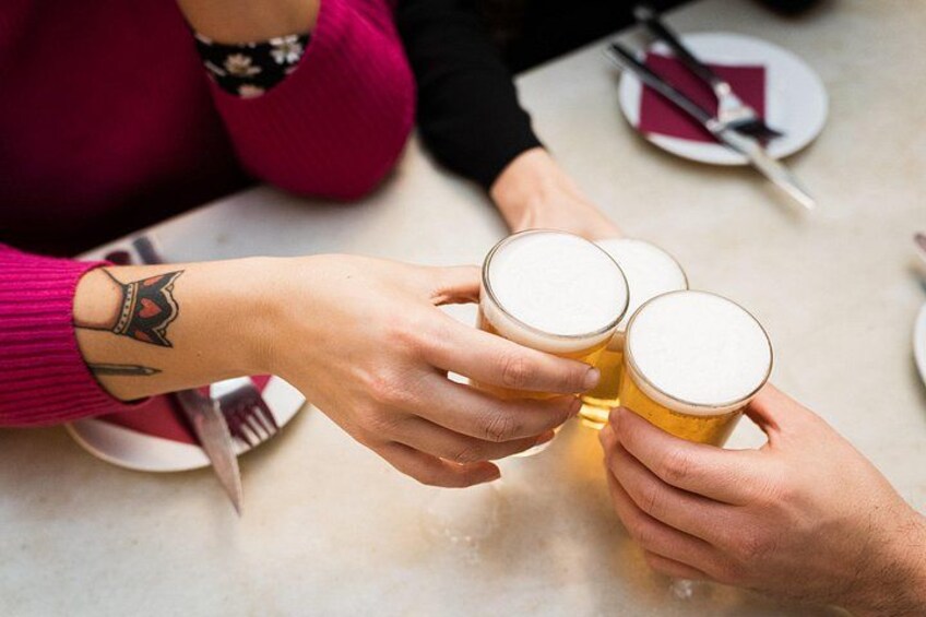 Tre hænder der holder ølglas, et bord med tallerkener og bestik, og en person med en tatovering på armen. 3