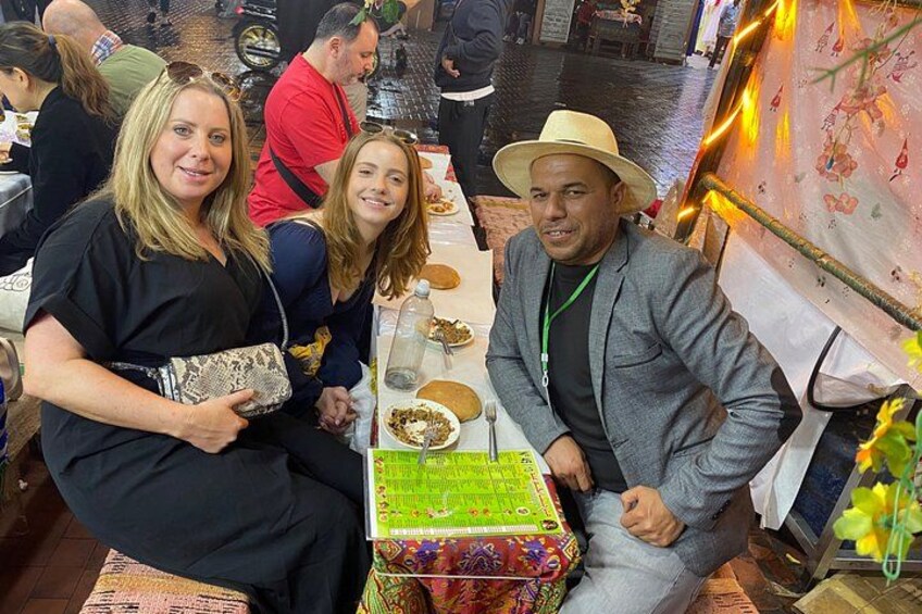 A group of people seated at a table with a cake and a floral tablecloth. 1