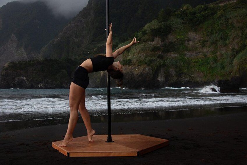 A person performing a pole dance on a wooden platform by the sea with a mountainous backdrop. 4