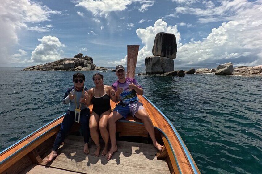 Three people on a wooden boat in the ocean with a large rock formation in the background. 1