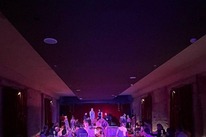 Een podium waar een band optreedt, een publiek zit aan tafels, en een kamer in gedimd licht. 3