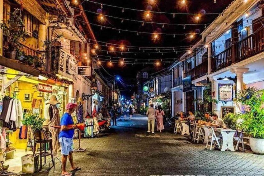 A nighttime street scene with shops and outdoor seating, string lights overhead, and a cobblestone street. 4