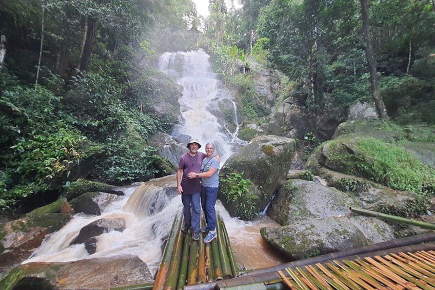 Una cascada en un área boscosa con una pareja de pie sobre un puente de bambú. 5