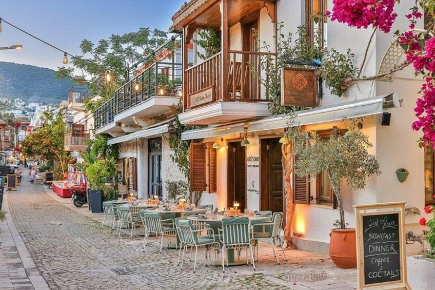 Una calle adoquinada bordeada de mesas al aire libre en un restaurante, con un cartel que anuncia desayuno, cena, café y cócteles. 2