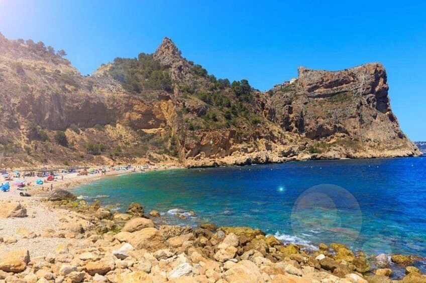 A rocky beach with clear blue water, a rocky cliff, and a clear sky. 5