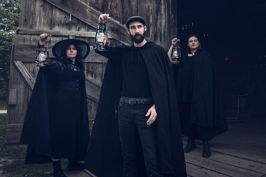 Three people in black attire holding lanterns, standing in front of a wooden structure. 1