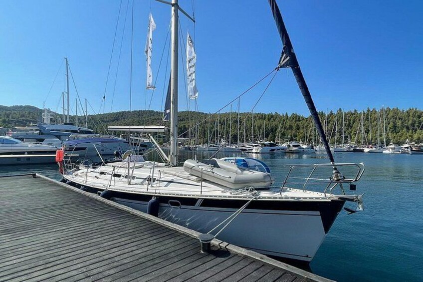 Een zeilboot afgemeerd in een jachthaven, met andere boten op de achtergrond, een houten steiger en een heldere blauwe lucht. 2