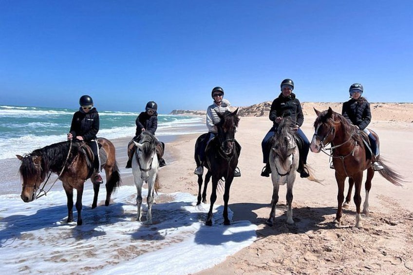 Un groupe de personnes à cheval sur une plage, avec l’océan en arrière-plan. 1