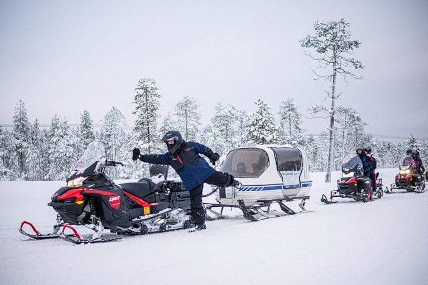 Una moto de nieve con una persona de pie a su lado, un remolque para motos de nieve y otra moto de nieve al fondo. 1