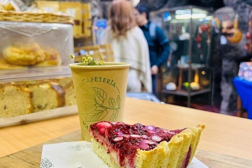 A slice of cake with a red fruit topping on a plate, a coffee cup with 'CAFETERIA' written on it, and a display case with pastries in the background. 3