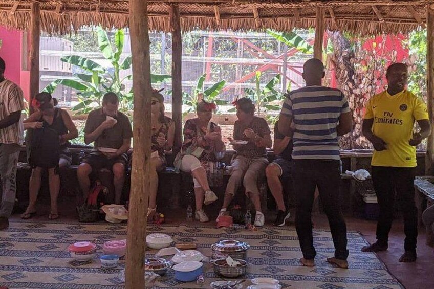 A group of people sitting on benches under a thatched roof, with various bowls and dishes on the floor. 2