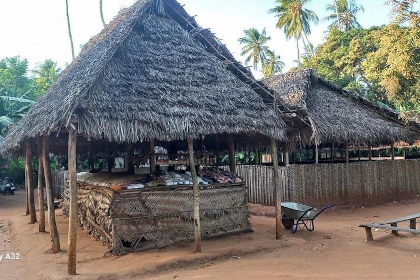 A thatched-roof hut with a woven bench and wheelbarrow, surrounded by palm trees. 4