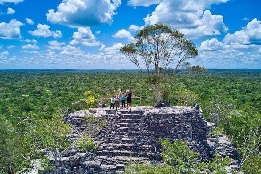 A stone structure with people standing on it, surrounded by lush greenery and a clear sky. 1
