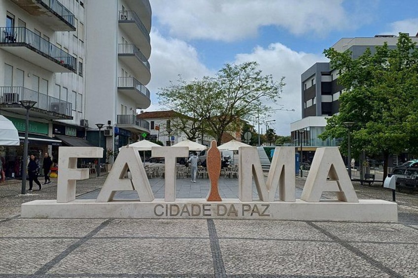 Un letrero con la inscripción 'FATIMA CIDADE DA PAZ' en una plaza pública, con edificios y árboles al fondo. 4