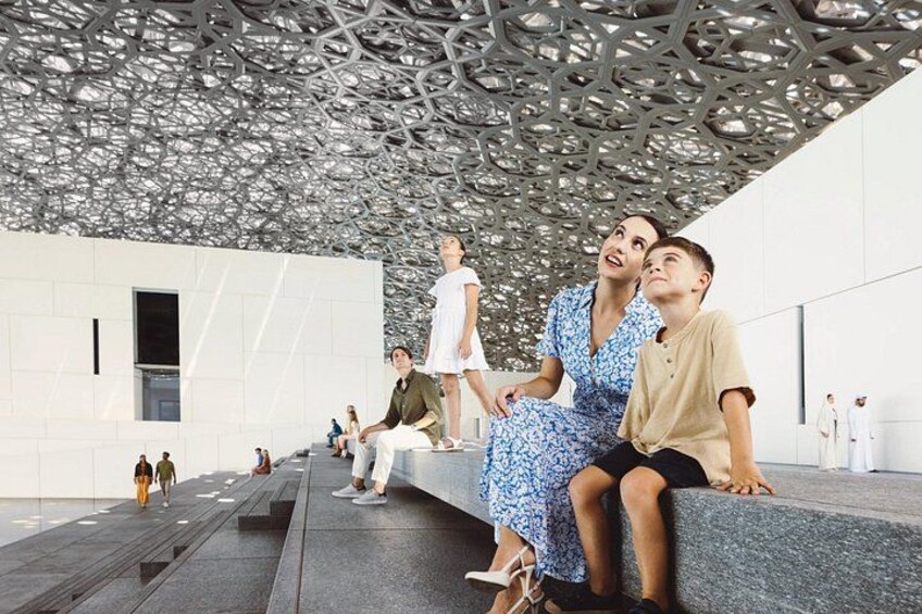Una estructura arquitectónica moderna con un techo geométrico y un grupo de personas sentadas en escalones. 1