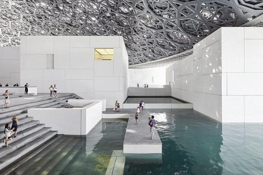 Un espacio arquitectónico moderno con techo geométrico, una fuente central y una serie de escalones que conducen a una plataforma. 3