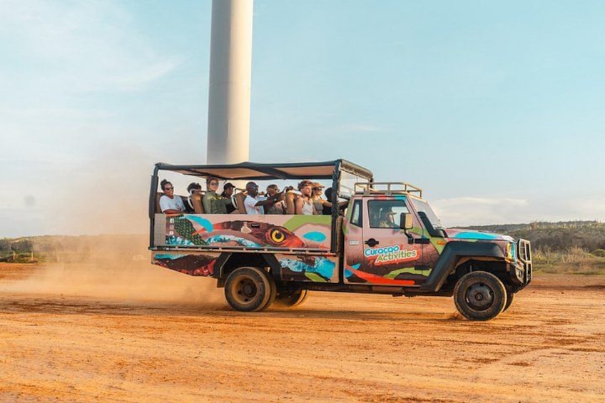 Un camión colorido con pasajeros circulando por un camino de tierra, con una gran turbina eólica al fondo. 5