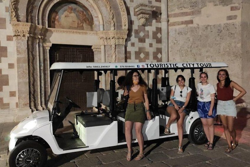 Un véhicule de golf blanc, portant une pancarte indiquant « VISITE TOURISTIQUE DE LA VILLE », est garé devant un immeuble à la façade carrelée et à l’entrée en arc de cercle. 1