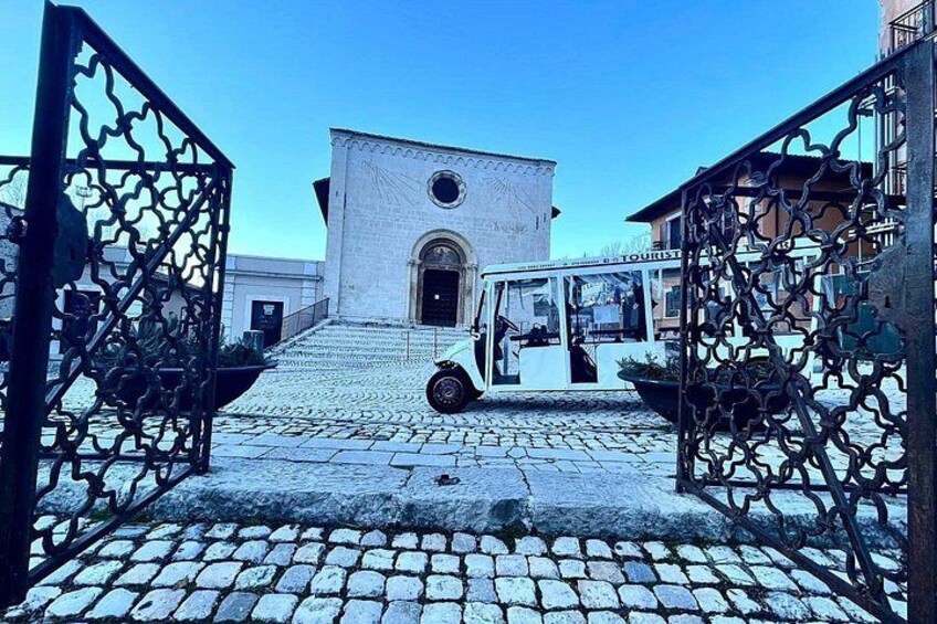 Une rue pavée menant à un bâtiment blanc surmonté d’une tour horlogère, avec un autocar blanc garé devant et de somptueuses grilles en métal. 4